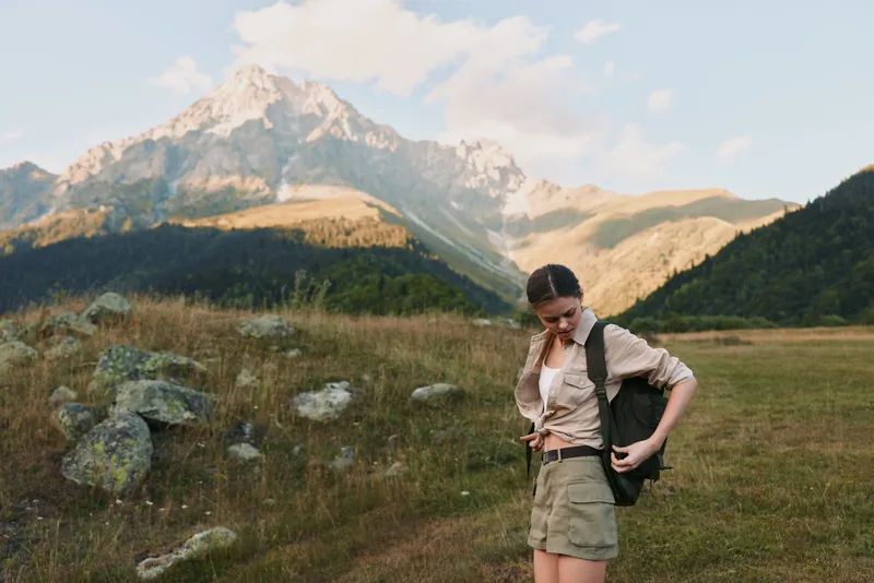 Young woman adjusts her backpack in a mountain meadow, smiling slightly as rugged peaks rise in the distance.