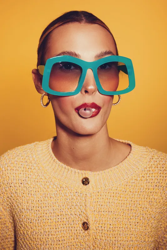 Young woman with oversized blue sunglasses and lip piercing poses against yellow background wearing knitted sweater. Concept of fashion, style, accessories, and modern attitude.