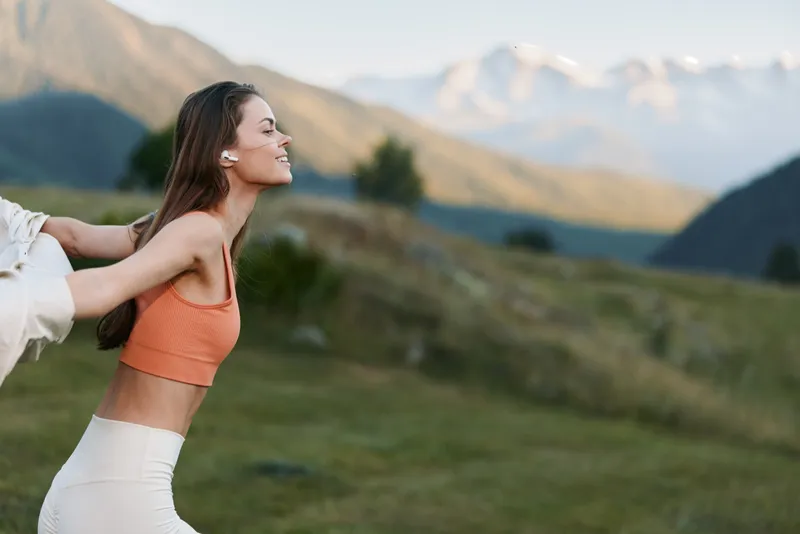A joyful woman outdoors in a scenic mountain landscape wears an orange top and white pants, smiling as she leans forward while a partner gently holds her hands.