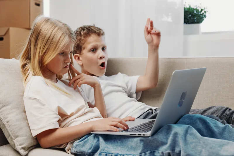 Two children sitting on sofa using laptop together with surprised and focused expressions in bright living room. Concept of technology, learning, and childhood interaction.