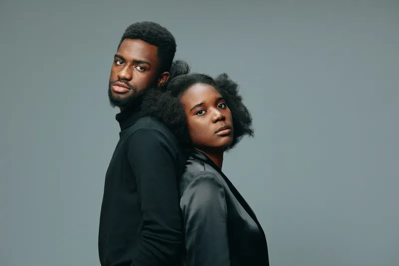 A striking portrait of a confident Black couple standing back to back, showcasing strong emotions and modern fashion in a minimalist studio setting