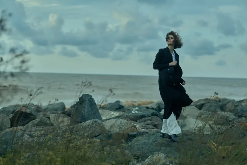 Woman in a long coat stands on a rocky shoreline with a windswept dress, gray clouds overhead and the sea in the distance, creating a moody cinematic landscape.