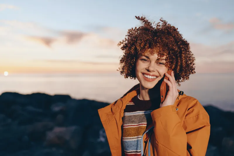 Woman, curly, hair, smiling, outdoors, lifestyle portrait, sunset, jacket, touching her ear, orange outerwear, cheerful expression, mountain landscape, warm sky background, casual travel mood
