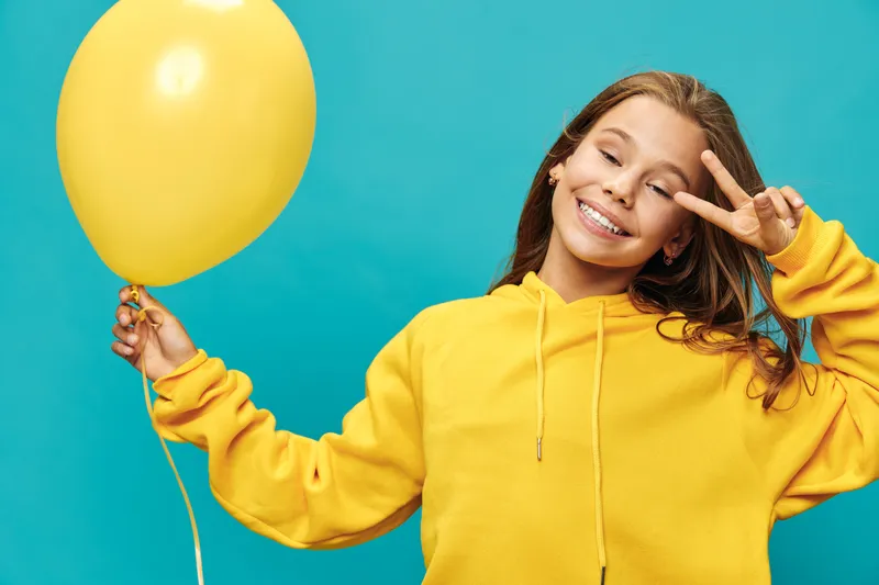 Girl smiling and holding yellow balloon with hand gesture on blue background. Casual clothing, happy child portrait, isolated with cheerful face and playful mood in studio shot.