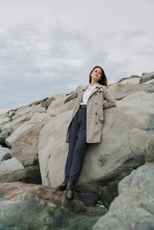 Young woman in casual coat leans against large rock on rocky shore under cloudy sky. Relaxed pose and natural setting create mood of calm and contemplation by the sea.