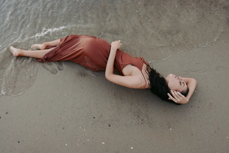 A woman in a flowing red dress rests on a wet sandy beach, with gentle waves around her, creating a calm, contemplative mood and a soft, romantic portrait feel.