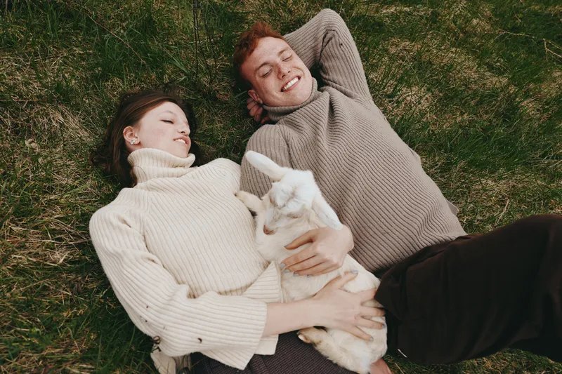 Happy couple relaxing on grass with a playful goat kid, enjoying outdoor lifestyle moments, cozy sweaters, gentle smiles, candid affection, intimate everyday joy, natural light
