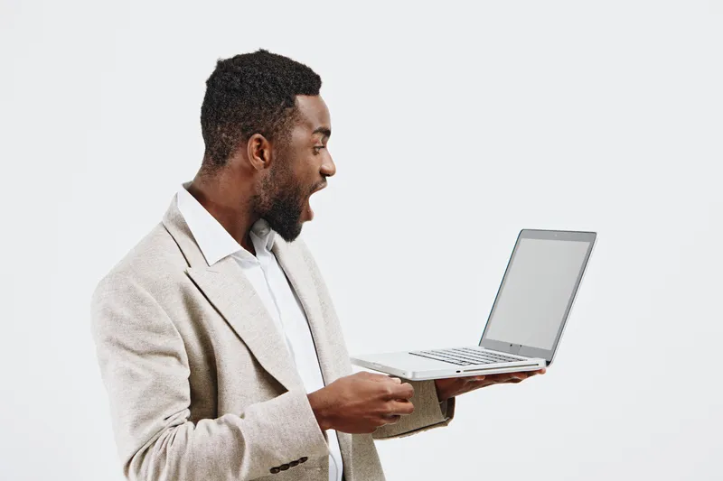 Man with laptop wearing beige blazer, excited and surprised, holding open notebook computer on white background, studio portrait. Business and technology concept