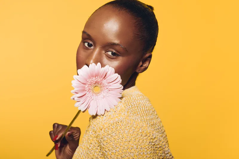 Portrait of a dark skinned African woman holding a pink flower near her face against a yellow background. Concept of beauty, natural skin, floral, and happiness in studio lighting.