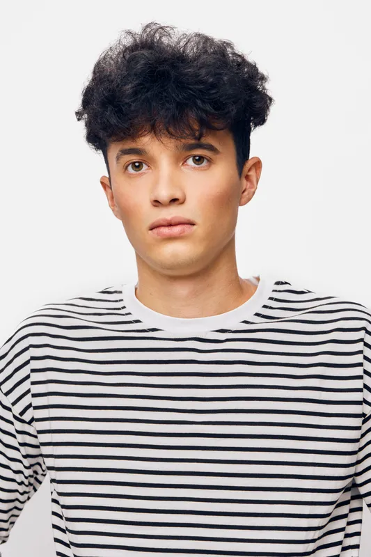 Young man with curly hair wearing a black and white striped shirt, posing against a plain white background, displaying a neutral expression and casual style.