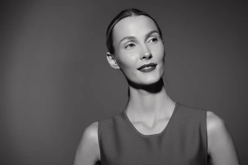 Portrait of a fair skinned Caucasian woman with slicked back hair in sleeveless dress looking away. Studio black and white lighting emphasizes elegant confident mood.