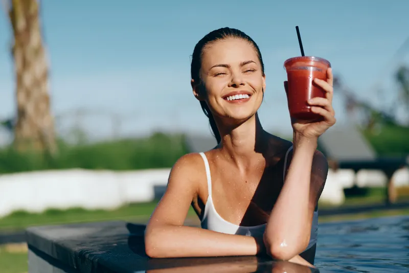 Smiling woman rests on the pool edge, enjoying a refreshing smoothie by the water, capturing a casual outdoor summer lifestyle, wellness and hydration in a sunlit moment.