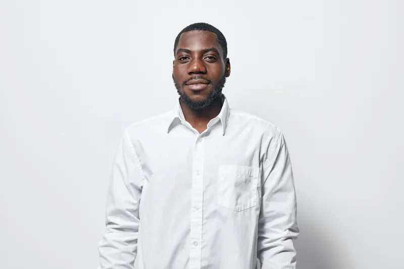 African American man with dark skin wearing white shirt standing against white background looking at camera with confident expression and relaxed posture in studio.