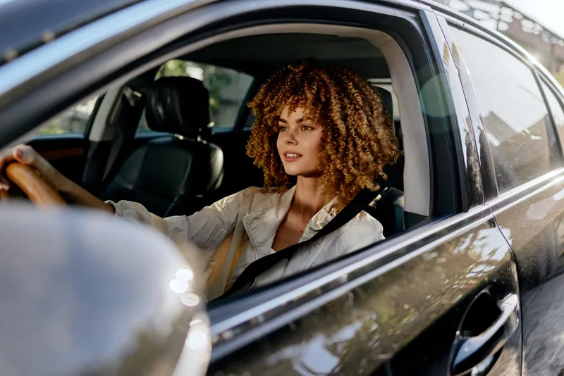 Young woman with curly hair driving car, wearing seatbelt and light jacket, focused and relaxed inside modern vehicle during daytime.