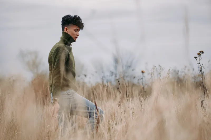 Young man walking through tall dry grass in a wool sweater, captured with natural light and a candid composition that emphasizes authenticity and reliability in a quiet outdoor portrait.