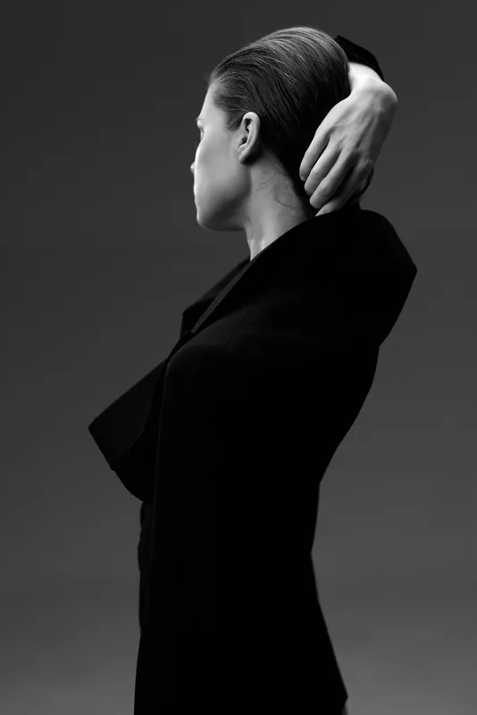 Black and white portrait of a woman in a black jacket posing with hand behind head, showcasing elegance and style in a minimalistic setting.