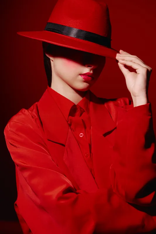 Elegant woman in red fashion outfit and red hat partially covering face on red background. High contrast lighting emphasizes stylish, creative beauty and fashion concept.