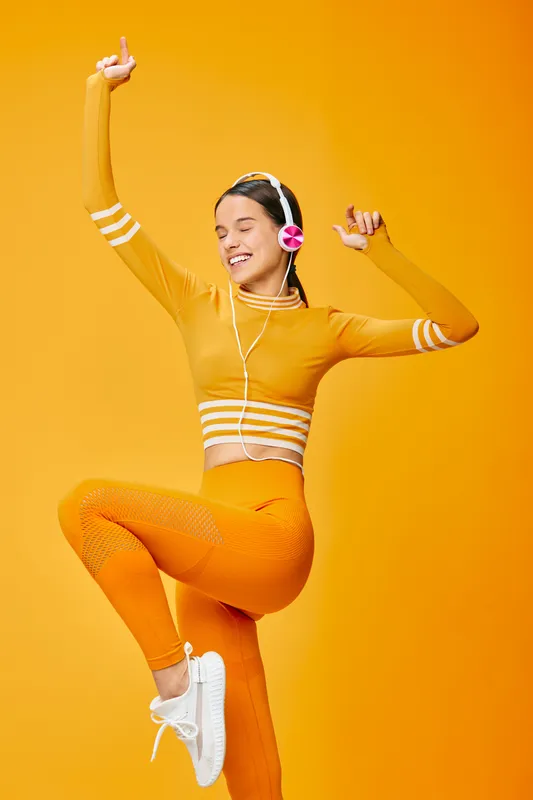 Woman fitness dancer in orange outfit jumps with energy against a vibrant orange background, wearing headphones, showcasing motion, active performance and studio style.