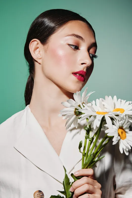 Young woman with bright makeup and glossy lips holding white daisies in a white blazer. Fashion beauty concept with soft studio lighting and a pastel green background.