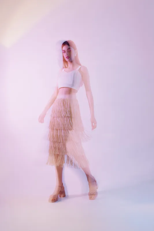 Young woman in a white crop top and beige fringed skirt poses against a soft purple background. Concept of woman beauty fashion, elegance, and style.