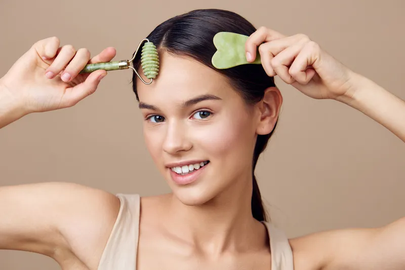 Young woman using jade roller for facial massage and skin care, showcasing healthy beauty routine and self care, against a soft beige background