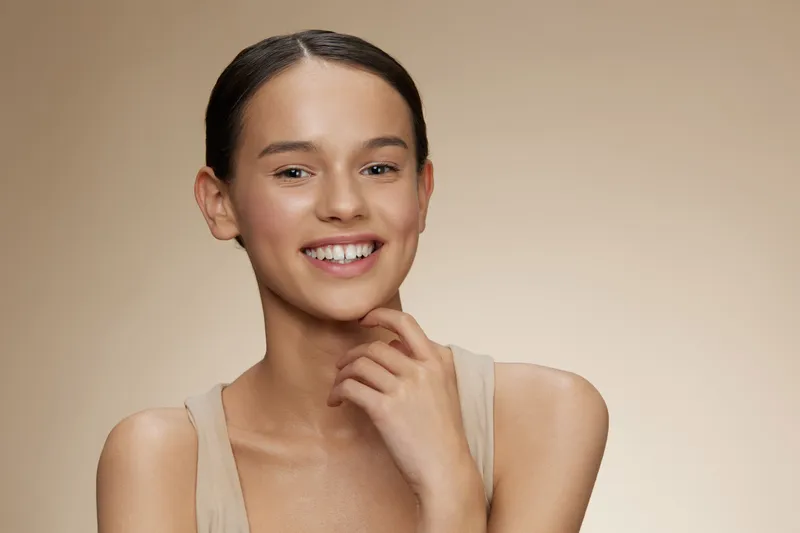 Portrait of a joyful young woman with dark hair, wearing a light beige top, smiling confidently against a smooth beige background, highlighting beauty and self assurance
