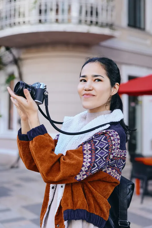 Woman photographer holding a camera outdoors on a sunny street, capturing moments with a colorful knit jacket, urban backdrop, candid lifestyle and creative focus