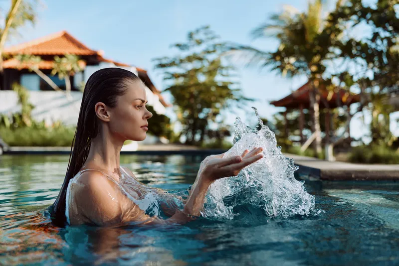 Young woman with braided hair enjoys splashing water while standing in a swimming pool at a sunny resort with tropical trees and buildings in the background.