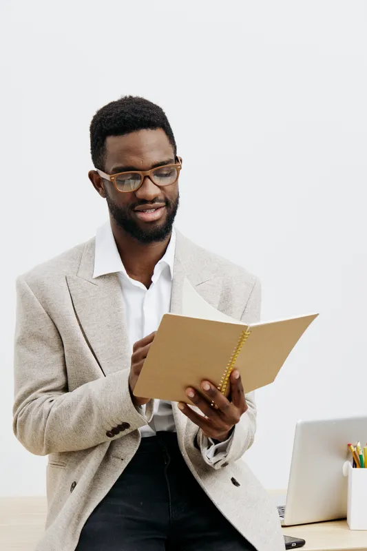 Confident smiling man of African descent wearing glasses, beige blazer, white shirt, and dark pants reading a yellow notepad, use of workspace setting with desk and laptop isolated on white background