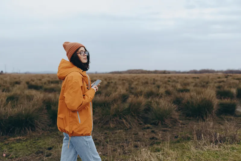 Woman lifestyle outdoors in autumn field, wearing yellow coat, walking through tall grass, holding notebook, thoughtful mood, cloudy sky background, calm nature day on countryside trail