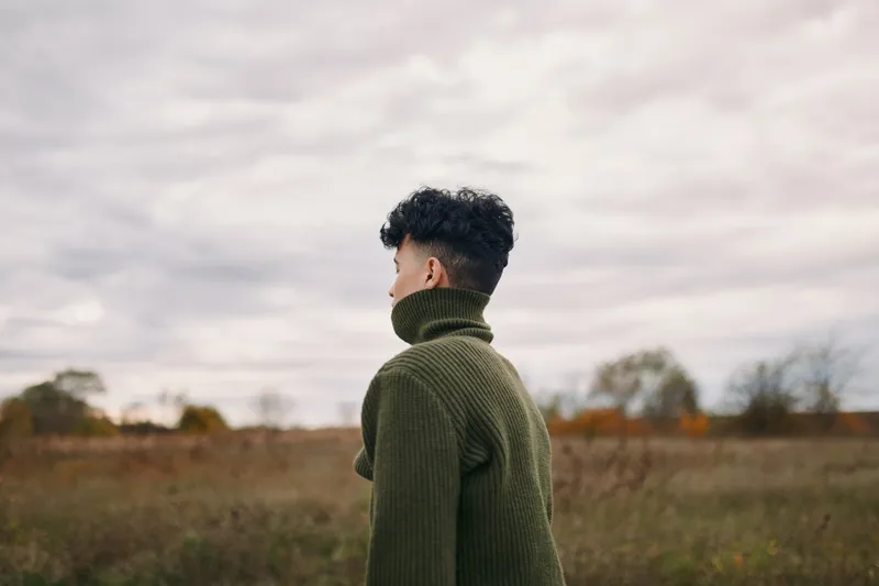 A candid image emphasizes authenticity as a single figure in a green knit turtleneck stands in a wide field, back turned, inviting honest interpretation and quiet focus.