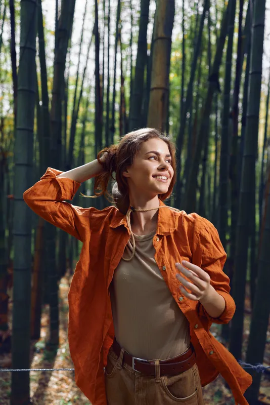 Woman bamboo forest portrait smiling in orange jacket, nature outdoors travel scene with happiness and sunlight. Casual style, relaxed pose, eco tourism and joyful outdoor lifestyle.