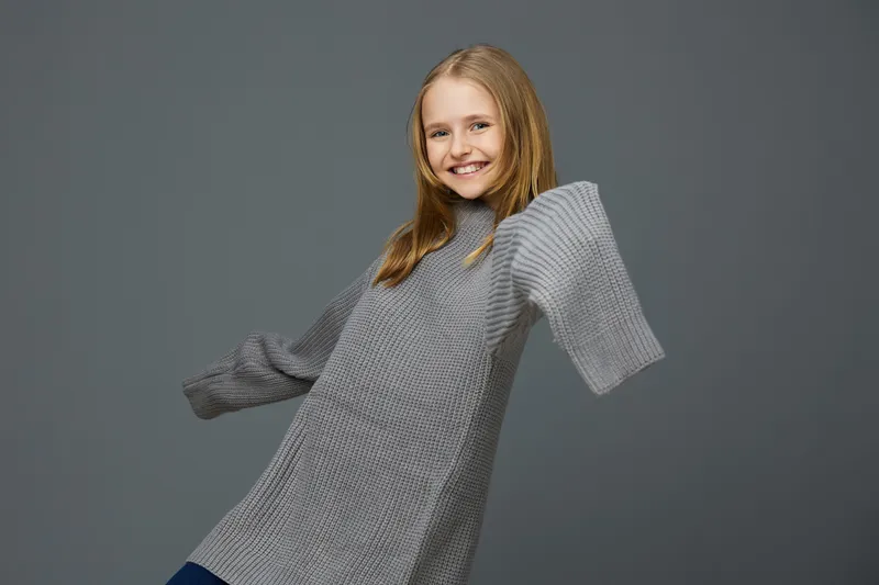 Young girl smiling in a cozy oversized gray sweater, joyful expression against a soft gray background, perfect for seasonal fashion and lifestyle themes