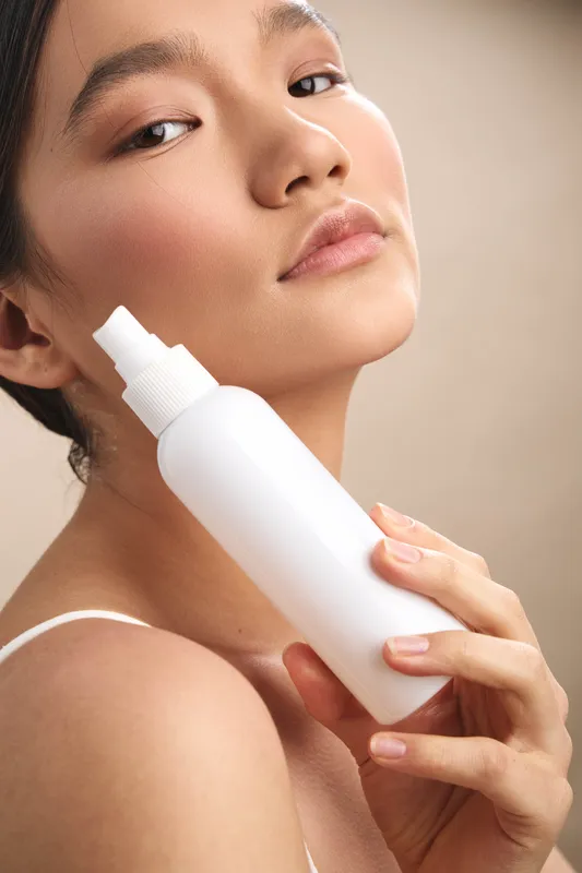 Asian woman applies skincare from a white bottle in a closeup studio shot, conveying calm concentration and a healthy glow on smooth skin in a modern setting.
