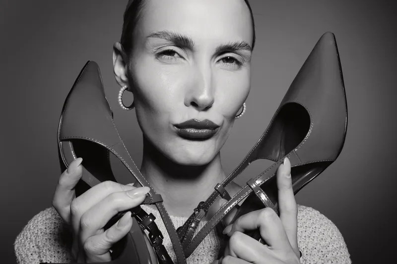 White woman with pale skin holds high heel shoes near face, posing with confident expression. Concept for fashion, beauty, and footwear style.