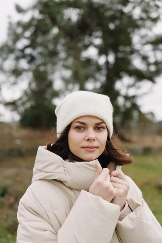 Woman in a beige winter coat and white beanie stands outdoors in a misty park, calm expression, cozy fashion detail for seasonal apparel and outdoor lifestyle