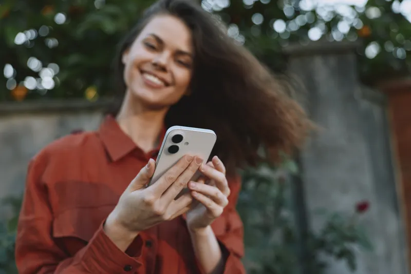 Young woman with brunette hair smiles while taking a selfie outdoors in a casual rust jacket, relaxed pose, soft natural light and leafy background