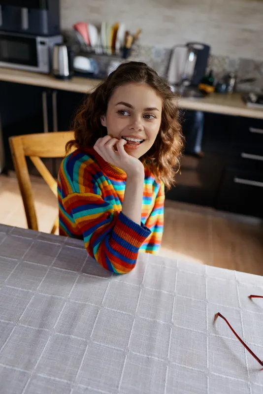 Young woman sits at a kitchen table, resting her chin on her hand and smiling softly, wearing a colorful striped sweater in a warm, homey, daylight kitchen scene.