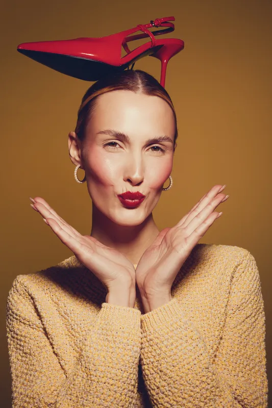 Young woman with red high heel shoes on her head poses with hands near face, wearing yellow sweater and gold earrings. Studio portrait on brown background with playful expression.