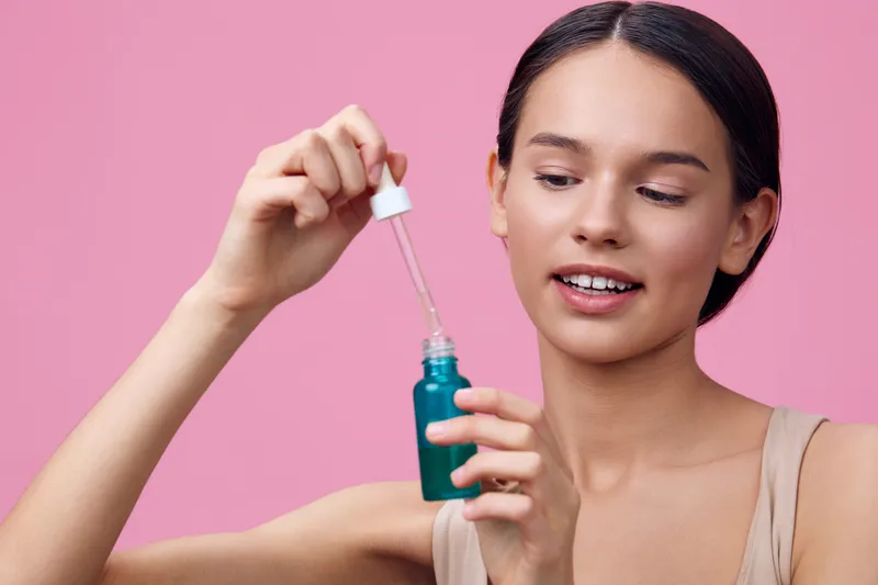 Young woman applying facial serum, showcasing healthy skin, smiling against a pink background, focusing on skincare and wellness routines