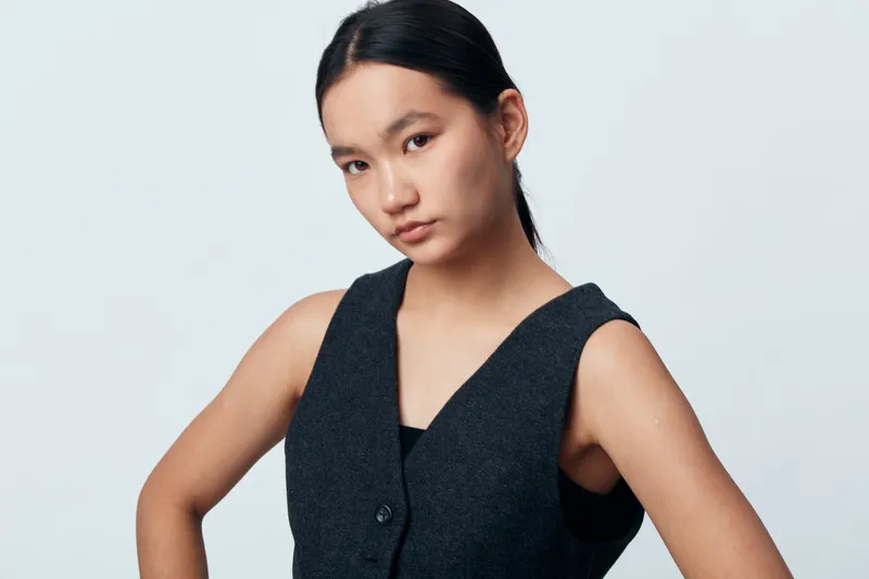 Young woman with serious expression posing in sleeveless dark vest on light background. Portrait, confident look, casual style, fashion, studio shot with natural lighting and minimal makeup.
