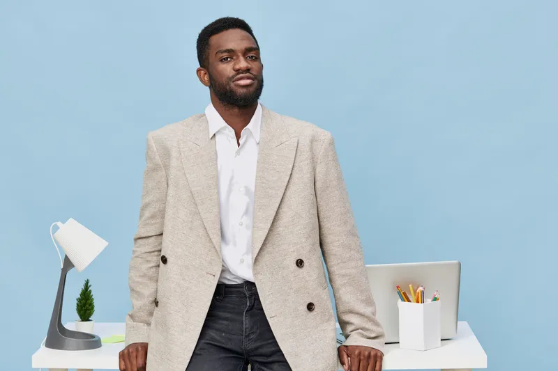 Confident young African American man in beige blazer and white shirt standing behind desk with laptop and stationery on office background, professional businessman expression, modern workspace, lifest