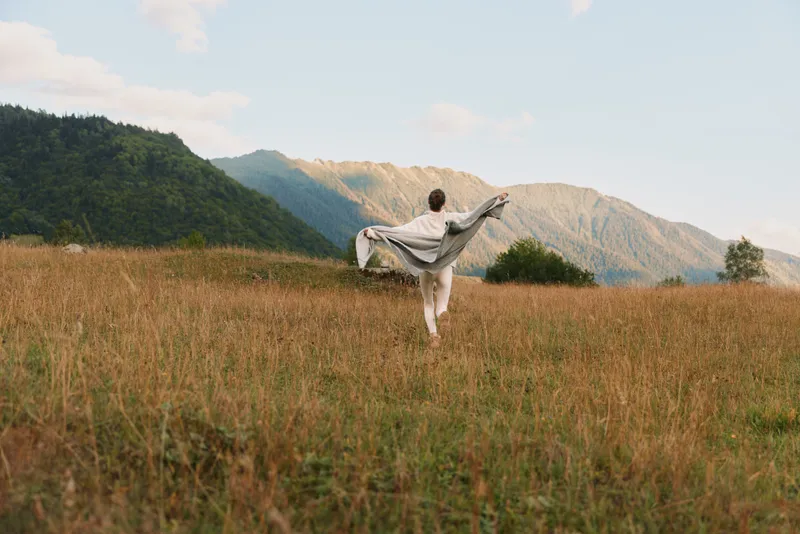 Young woman dances in a golden field with a flowing shawl, mountains rising behind her, radiating freedom and calm in late afternoon light, her dress moving with the breeze