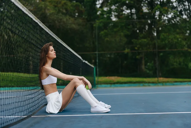 Fit young woman in white sportswear sitting on a tennis court, exuding confidence and determination, perfect for fitness and wellness concepts