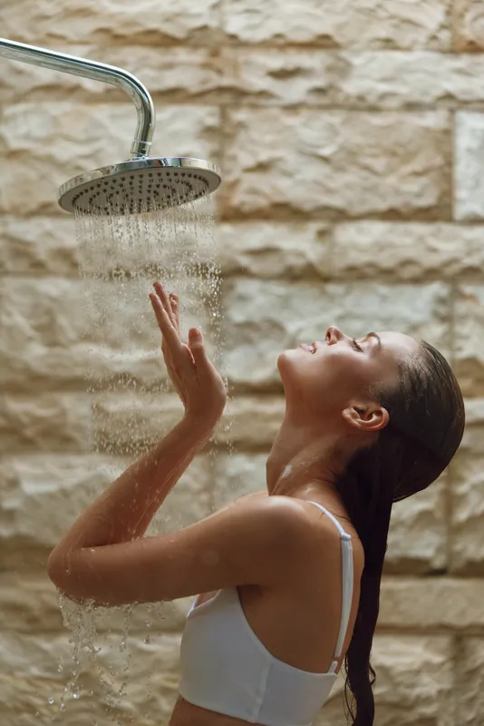 young woman enjoying water shower outdoors with relaxed expression, wet hair, wearing white top, natural stone wall background, refreshing moment