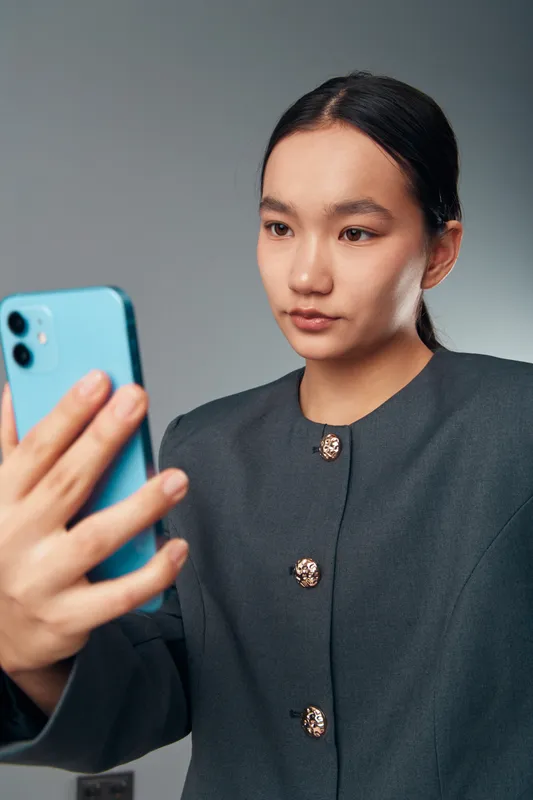 Young woman in dark jacket holding smartphone looking at screen with focused expression on neutral background. Female using mobile phone for video call or selfie indoors.