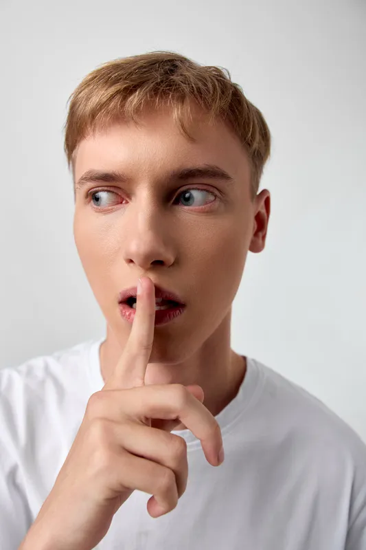 Shushing gesture by a young adult, finger pressed to lips, signaling secrecy and silence in a clean studio setting, minimal background, calm expression, suitable for messaging about discretion,