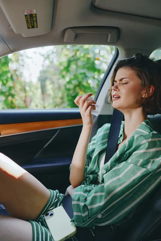 A woman rides in a car, appearing unwell and nauseous as she holds a small object near her mouth during motion.
