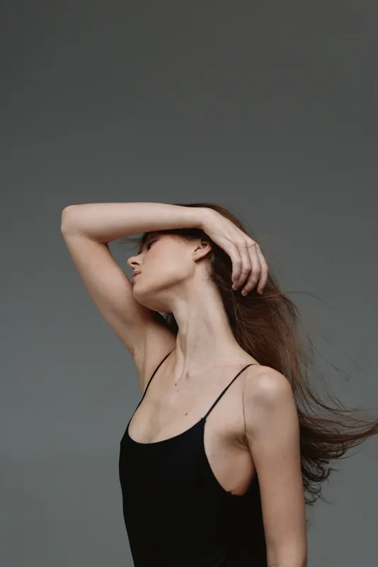 Elegant woman in black top posing with hand on head against neutral background, showcasing graceful movement and natural beauty.