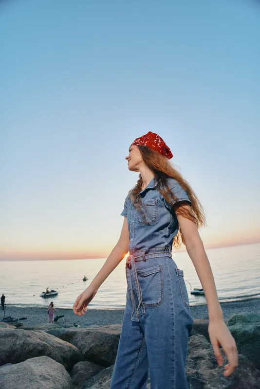 Lifestyle woman in modern boho-western denim street style stands by the sea during sunset, with film color effect enhancing relaxed and stylish mood.